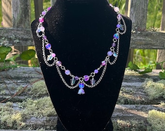 May include: A delicate silver chain necklace with iridescent purple and pink beads and flower charms. The necklace is layered with two chains and has a delicate, feminine aesthetic.