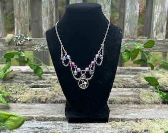 May include: A silver chain necklace with a charm of a mushroom and a small figure standing under it. The necklace has iridescent purple and blue beads and star-shaped charms.
