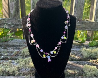 May include: A delicate necklace with pink, purple, and clear beads, and silver chain accents. The necklace features heart-shaped and star-shaped charms, and is displayed on a black velvet bust against a natural outdoor backdrop.