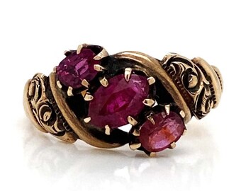 May include: A gold-toned ring featuring three oval-cut, deep red gemstones. The ring has an ornate, swirling design with detailed scrollwork on the sides. The gemstones are set in a prong setting. The ring is on a white background.