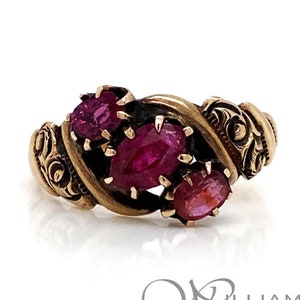 May include: A gold-toned ring featuring three oval-cut, deep red gemstones. The ring has an ornate, swirling design with detailed scrollwork on the sides. The gemstones are set in a prong setting. The ring is on a white background.