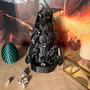 May include: A black 3D printed dragon tower with a dragon head on top. The tower is surrounded by smaller dragons and has a dice tray at the bottom. There are several dice scattered on a brown surface.