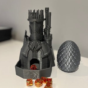 Peut inclure: Un château gris imprimé en 3D avec un œuf de dragon et des dés rouges et dorés. Le château a une tour avec un horizon de ville au sommet et une crête de dragon à l'avant. L'œuf de dragon est une sphère grise texturée.