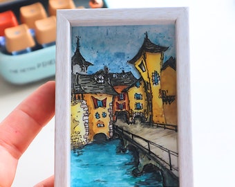 May include: Watercolor painting of a European town with yellow and orange buildings, a bridge, and a blue river. The artwork is in a white frame. The painting is held in a hand.