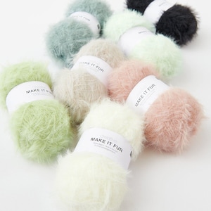 Può includere: Una collezione di gomitoli di lana soffice in vari colori pastello, tra cui verde, rosa e crema. Ogni gomitolo ha un'etichetta con la scritta "MAKE IT FUR". La lana appare morbida e strutturata, adatta per progetti di maglieria o uncinetto.