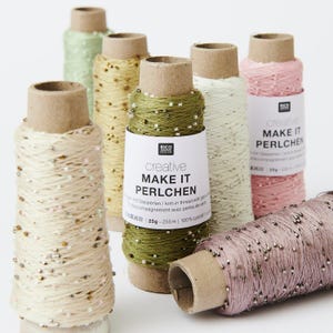 Könnte beinhalten: Verschiedene Garnspulen in Creme-, Grün- und Rosatönen. Das Garn ist um Pappröhren gewickelt und mit kleinen Perlen verziert. Auf dem Etikett steht "creative MAKE IT PERLCHEN".