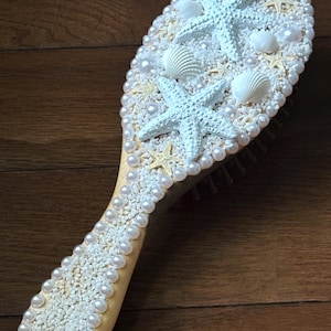 May include: A wooden hairbrush decorated with white seashells, starfish, and faux pearls. The brush head is covered in small white stones, with larger starfish and seashells. The handle is also decorated with pearls and stones.