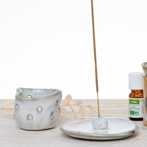 Può includere: Porta incenso in ceramica bianca con un top rotondo aperto e una superficie testurizzata e irregolare. Il porta incenso è su una superficie di legno con un unico bastoncino di incenso acceso al centro.
