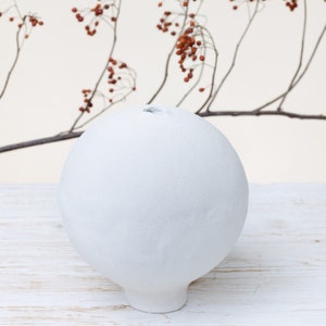 Può includere: Un vaso in ceramica bianca con una forma rotonda e bulbosa e una piccola apertura nella parte superiore. Il vaso è appoggiato su una superficie di legno bianca.