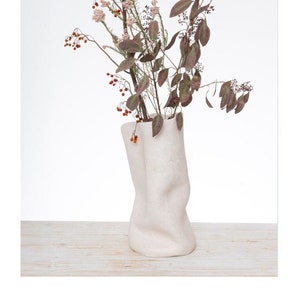 Può includere: Un vaso di ceramica bianca con una superficie testurizzata e stropicciata. Il vaso è pieno di fiori e bacche secche.