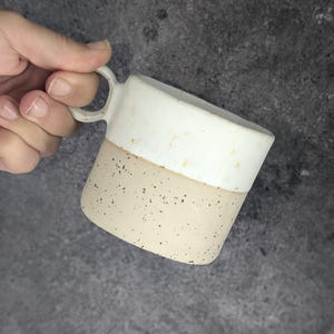 Può includere: Una tazza in ceramica bianca con un fondo beige macchiato. La tazza ha un manico arrotondato.