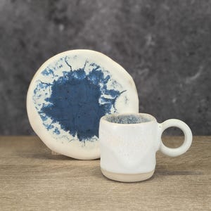 Può includere: Una tazza da caffè in ceramica bianca con un design a spruzzo blu e un piattino abbinato. La tazza ha una forma arrotondata e un piccolo manico.