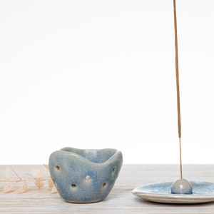 Può includere: Un porta incenso in ceramica blu con un piccolo foro rotondo al centro. Il porta incenso è appoggiato su una superficie di legno bianco. Un piatto per incenso in ceramica blu con un piccolo foro rotondo al centro è anche sulla superficie. Un bastoncino di incenso marrone sporge dal foro nel piatto.