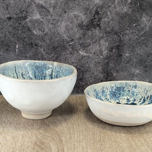 Può includere: Due ciotole in ceramica bianca con una glassa macchiata di blu e bianco. Le ciotole sono di dimensioni diverse e sono appoggiate su una superficie di legno.