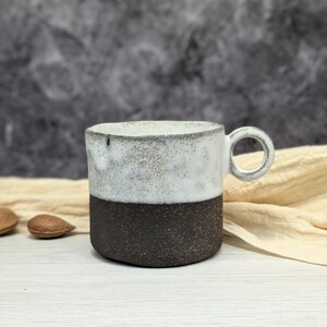Può includere: Una tazza in ceramica bianca e nera con un manico arrotondato. La tazza ha una glassa bianca macchiata sulla metà superiore e una glassa marrone scuro sulla metà inferiore.