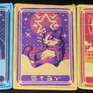 May include: Three rectangular embroidered patches featuring pixel art cat designs. The left patch is blue with a moon and the text Moon. The centre patch is yellow with a star and the text Star. The right patch is light yellow with a sun and the text Sun.