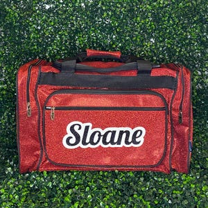 May include: A red glitter duffel bag with black trim and black webbing handles. The front features a zippered pocket with the name Sloane written in a bold, black, cursive-style font with a white outline. The bag has multiple black zippers.