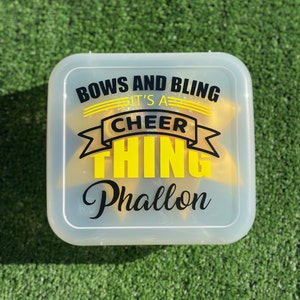 May include: A clear plastic container with a yellow and black design that says "Bows and Bling It's a Cheer Thing Phallon".