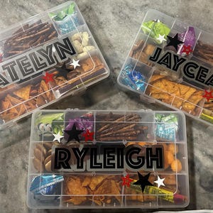 May include: Three clear plastic snack boxes, each divided into compartments and filled with various snacks like chips, pretzels, and candies. Each box is personalized with a name in bold black letters and decorated with star accents.