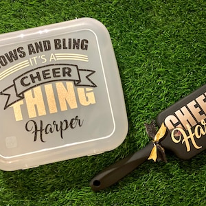 May include: A white plastic storage box with the text "Bows and Bling It's a Cheer Thing Harper" and a black hair brush with the text "Cheer Harper" in gold glitter lettering.