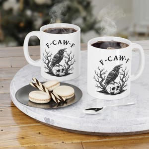 May include: Two white ceramic mugs filled with dark coffee, each featuring a black graphic of a raven perched on a human skull surrounded by bare trees and the text F-CAW-F. A small black plate holds three round biscuits and two striped wafer sticks.