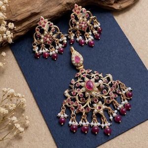 May include: A gold-coloured jewellery set comprising a pendant and matching earrings. The set is decorated with ruby-coloured stones and small white pearls. The pendant and earrings have an ornate, floral design.