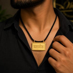 May include: A gold rectangular pendant featuring the Punjabi text "ਬਲਜੀਤ" in raised lettering, suspended from a black braided cord. The pendant has a textured border and a polished finish, hanging against a black collared shirt.