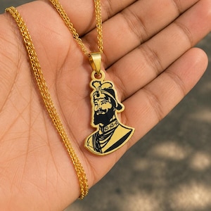 Può includere: Una collana con ciondolo in metallo color oro caratterizzata da una dettagliata silhouette nera incisa di una figura barbuta che indossa un turbante tradizionale e un abbigliamento ornato, appesa a una delicata catena dorata.