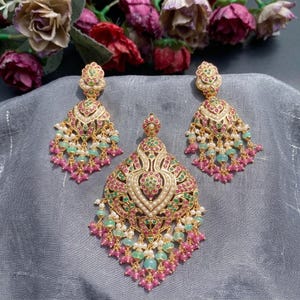May include: A gold-toned jewellery set consisting of a large pendant and two matching drop earrings. Each piece features intricate metalwork inlaid with small pink and green stones, accented by tiny white pearls and dangling bead clusters.
