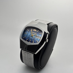 Peut inclure: Une montre Orient argentée avec un cadran bleu et un bracelet en maille. La montre a un boîtier rectangulaire avec un design angulaire unique. Le cadran affiche le jour et la date. La montre est présentée sur un support noir.
