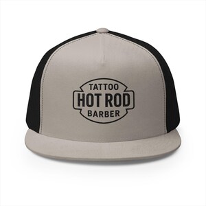 May include: A flat-peak trucker cap with a beige front panel and a black mesh back. The cap displays the text "Tattoo Hot Rod Barber" in a black, stylised font.