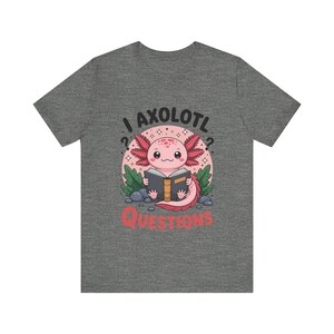 May include: A dark heather gray t-shirt featuring a graphic of a pink axolotl reading a book. The text above reads I AXOLOTL and the text below reads QUESTIONS. The design includes green leaves, small gray rocks, and white sparkles on a pink circle.