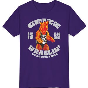 May include: Purple t-shirt with a graphic of a bear wearing a wrestling singlet. The bear graphic has the text "TRY ME" on the singlet. The shirt also has the text "GRIZZ WRASLIN' YELLOWSTONE" and "18 OR OLDER".