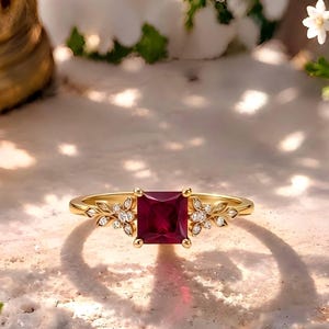 Könnte beinhalten: Ein Goldring mit einem quadratisch geschliffenen, tiefroten Edelstein in der Mitte, flankiert von kleinen blattförmigen Akzenten, die mit winzigen klaren Steinen besetzt sind, auf einer strukturierten, hellen Oberfläche.