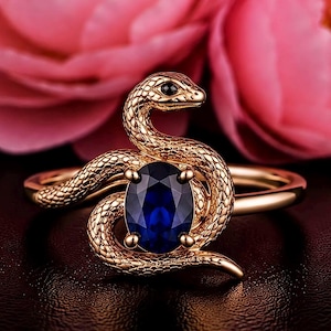 May include: A gold-toned ring featuring a coiled snake design with a textured, scale-like surface. The snake's body wraps around a central, oval-shaped, deep blue gemstone held in place by four small prongs. The snake has a small black eye.