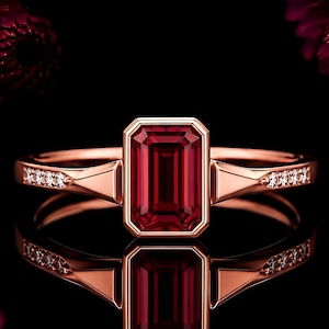 Könnte beinhalten: Ein Ring aus Roségold mit einem zentralen, smaragdgeschliffenen, tiefroten Edelstein in einer Zargenfassung. Die Ringschiene ist an beiden Seiten mit kleinen, klaren, rund geschliffenen Steinen besetzt, die zum Mittelstein hin verlaufen.