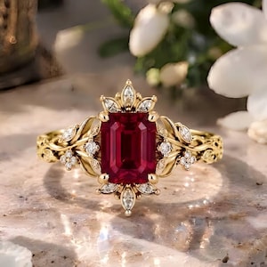 Puede incluir: Un anillo de oro con una piedra preciosa de color rojo intenso en talla esmeralda en el centro. La banda presenta un intrincado trabajo de metal en forma de hojas, adornado con pequeñas piedras claras en talla marquesa y redonda en patrón floral.