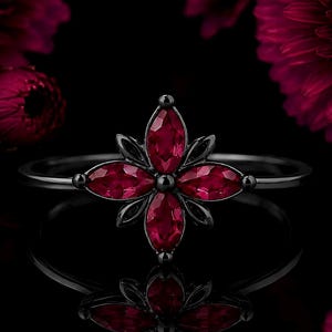 May include: A black metal ring featuring a central floral motif composed of four marquise-cut deep red gemstones arranged in a cross pattern, with smaller black metal accents between each stone, set on a thin, polished black metal band.
