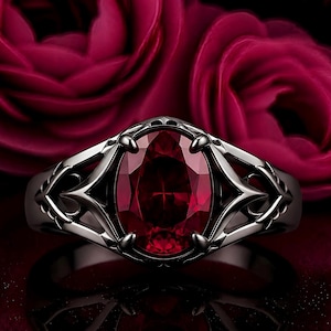 Puede incluir: Un anillo con una gran piedra preciosa de color rojo intenso y corte ovalado, engastada en una banda de tono gris oscuro. El trabajo en metal que rodea la piedra central muestra un diseño de celosía simétrico, puntiagudo y complejo.