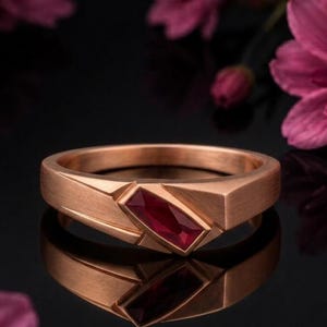 Peut inclure: Une bague en or rose ornée d'une pierre précieuse rubis rouge taille baguette rectangulaire sertie en diagonale sur l'anneau. L'anneau en métal présente une finition mate brossée avec des lignes géométriques nettes et angulaires.