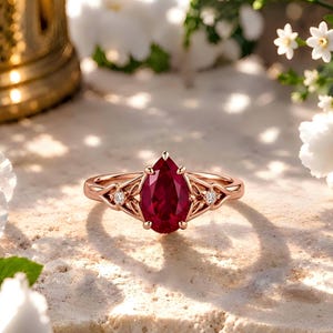 Può includere: Un anello in oro rosa con una gemma rosso scuro a forma di pera tenuta da griffe. La fascia presenta un'intricata lavorazione in metallo con piccole pietre rotonde trasparenti su ciascun lato dell'incastonatura centrale, creando un aspetto dettagliato.
