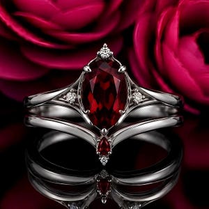 May include: A silver-toned ring featuring a large, oval-cut deep red gemstone as the centerpiece, accented by smaller clear stones. The band has a split design with a V-shaped contour, incorporating a small pear-cut red stone at the base point.