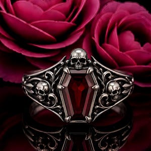 Puede incluir: Un anillo de plata con una piedra preciosa de color rojo intenso en forma de ataúd, flanqueado por dos calaveras detalladas a los lados y una calavera sobre el ataúd, con un fondo de intrincados adornos metálicos y pétalos de rosa oscuros.