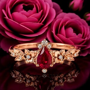Può includere: Un anello in oro rosa con una gemma rossa centrale a forma di pera, circondata da piccole pietre trasparenti e motivi floreali in metallo. La fascia è dettagliata con foglie scolpite e ulteriori piccole pietre trasparenti, su sfondo di rose.