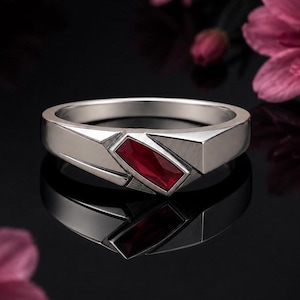 Peut inclure: Une bague en métal argenté ornée d'une pierre précieuse rectangulaire rouge foncé, taillée en baguette et sertie en diagonale sur l'anneau. L'anneau présente une finition polie lisse avec des détails géométriques angulaires autour du sertissage.