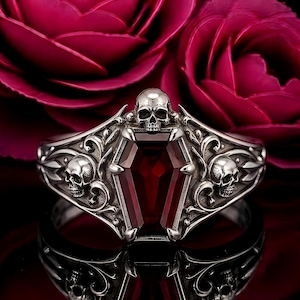 May include: A silver ring featuring a central coffin-shaped deep red gemstone. The band is decorated with intricate scrollwork and three detailed skull motifs, with one skull positioned above the coffin and two on the sides of the ring band.