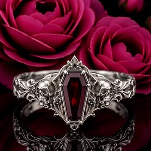 Peut inclure: Une bague en argent avec une pierre précieuse rouge foncé en forme de cercueil au centre, flanquée de deux motifs de crânes détaillés. L'anneau est orné de volutes complexes et de petites pierres noires, sur un fond sombre.