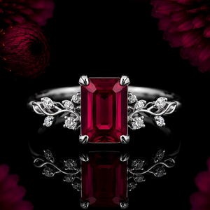 Könnte beinhalten: Ein silberner Ring mit einem großen, rechteckigen, smaragdgeschliffenen tiefroten Edelstein in der Mitte. Die Ringschiene ist auf beiden Seiten mit filigranen, rankenartigen Metallverzierungen gestaltet, die mit kleinen, runden, klaren weißen Steinen besetzt sind.
