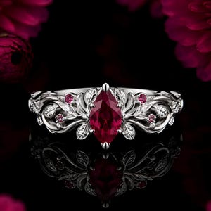 May include: A silver ring featuring a central marquise-cut deep red gemstone. The band is designed with intricate metal leaf motifs and small round red accent stones embedded along the sides, creating a detailed botanical appearance.