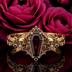 May include: A gold ring featuring a central coffin-shaped dark red gemstone, flanked by two detailed skull motifs on either side. The band is decorated with intricate scrollwork and accented with small, round, black gemstones.